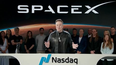 Speaker in a black leather jacket addresses a crowd at Nasdaq with a SpaceX banner and a line of team members behind him, celebrating the launch milestone.