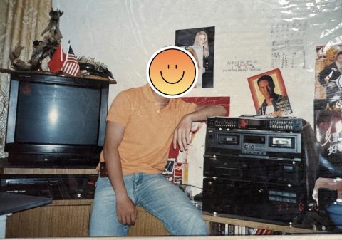 Person in an orange shirt and jeans sits next to a vintage stereo, with posters on the wall and an American flag nearby; face covered by a smiley sticker.